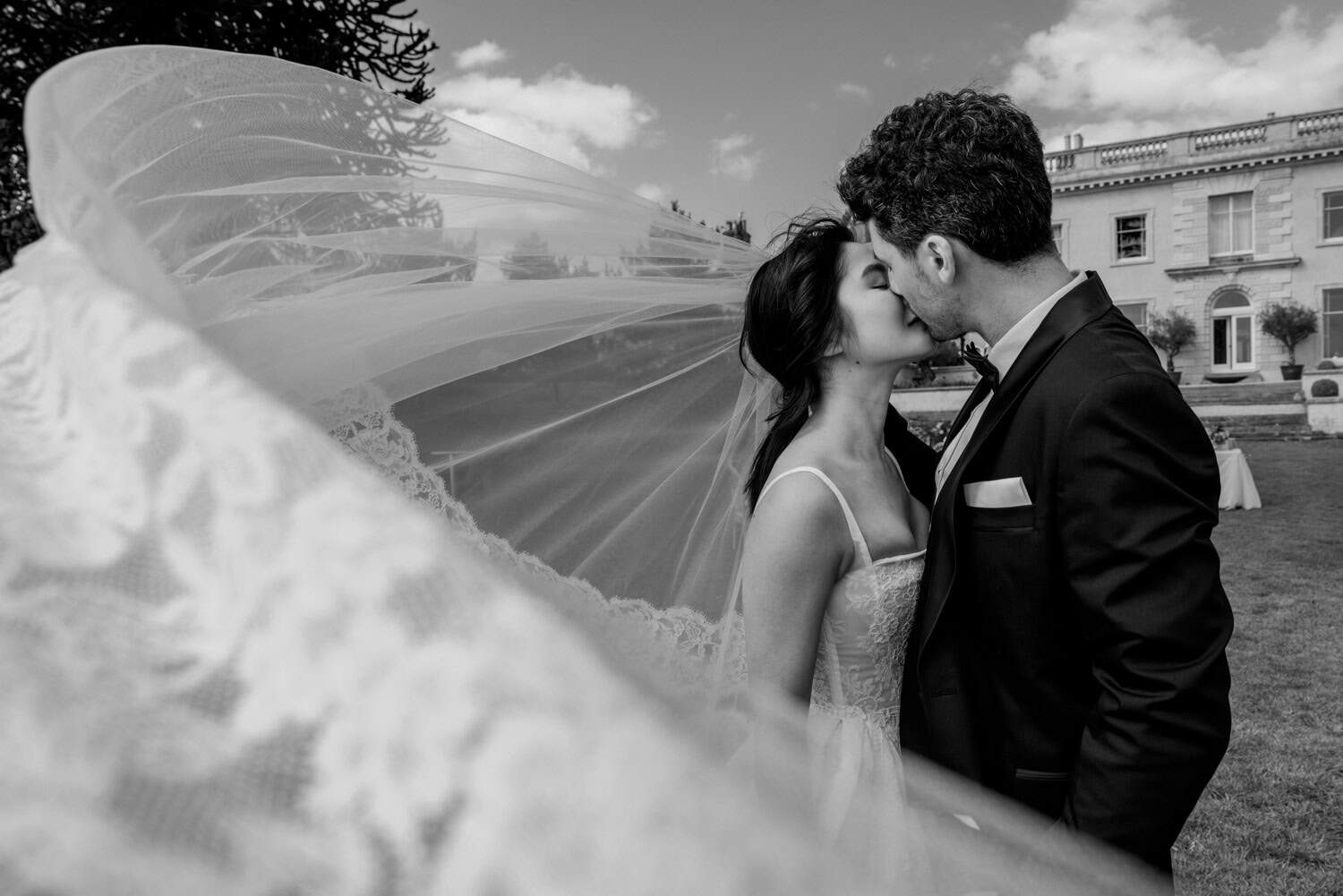 Bride and groom kissing outside The Location House as the bride’s veil blows dramatically across the foreground in a black and white wedding photograph.